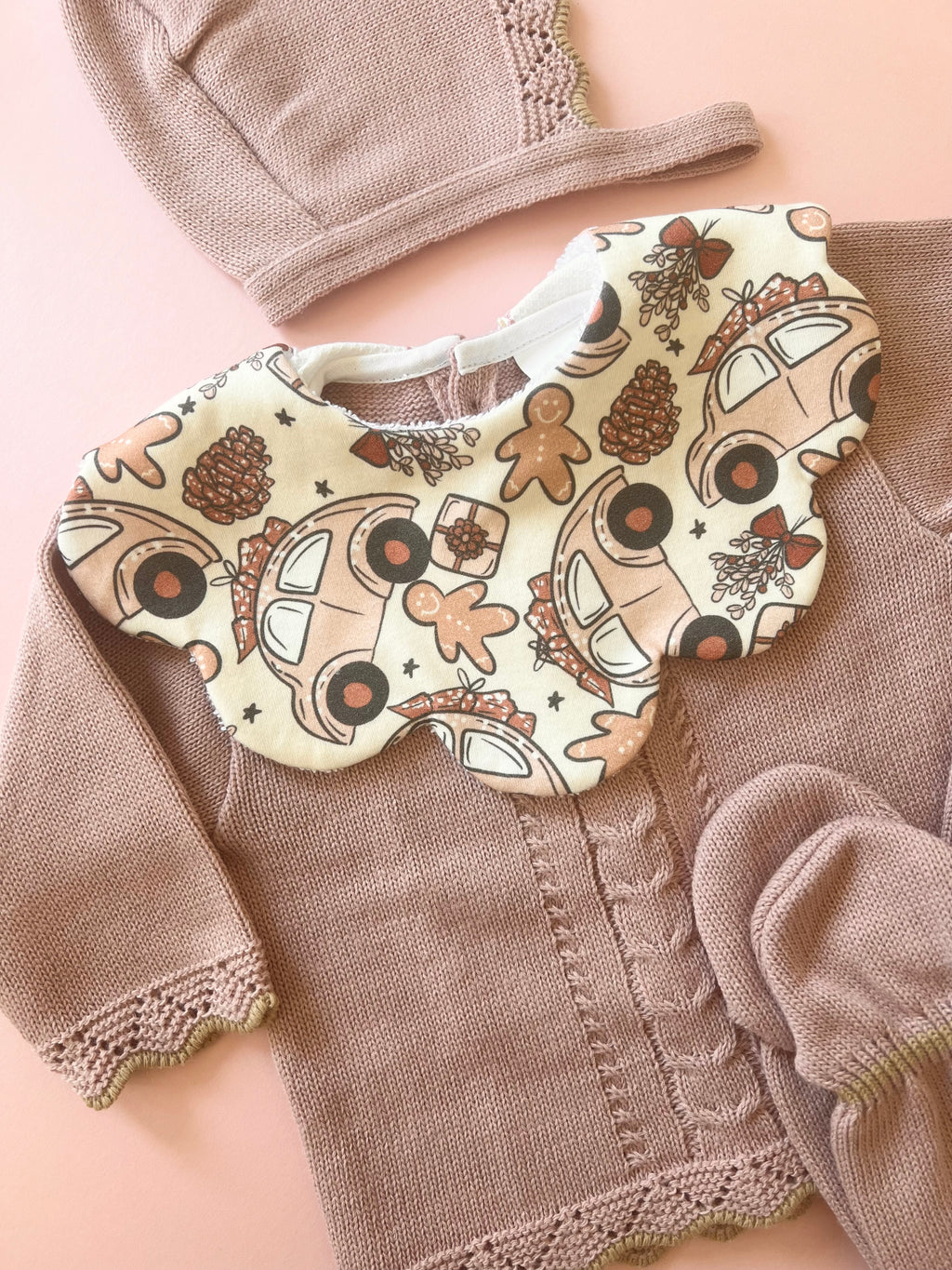 Scalloped baby bib in Liberty fabric with a festive print of cars, gingerbread men, and pinecones, paired with a blush pink knitted baby cardigan, bonnet, and mittens. A cosy winter outfit for babies, crafted with high-quality materials for style and comfort. Baby clothing set by Monica & Me.