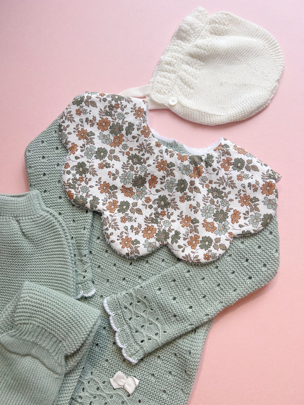 Scalloped baby bib in green and white floral Liberty fabric, styled with pastel green knit baby clothing featuring delicate lace edging and a cream knitted bonnet, set against a soft pink background.