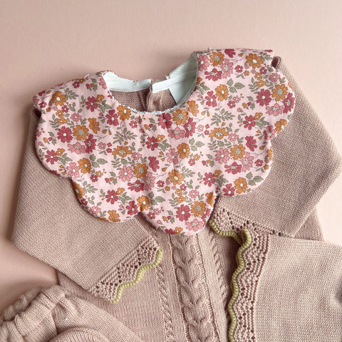 Delicate scalloped baby bib in pink floral Liberty fabric with vintage-inspired flowers, paired with soft neutral-toned knitted baby clothing featuring cable knit and scalloped edging, set against a warm beige background.