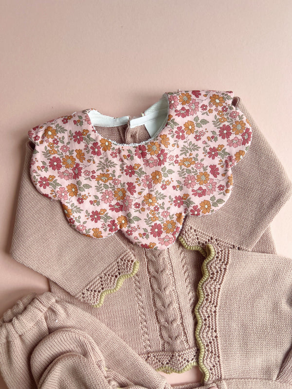 Delicate scalloped baby bib in pink floral Liberty fabric with vintage-inspired flowers, paired with soft neutral-toned knitted baby clothing featuring cable knit and scalloped edging, set against a warm beige background.